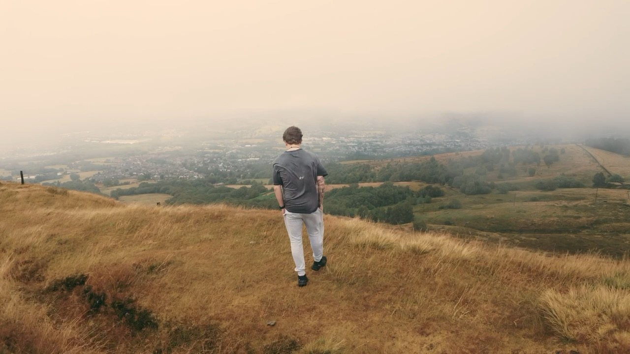 A person walking on a moorland hilltop, looking out over a hazy valley town.