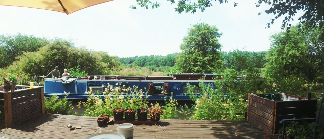 A blue narrowboat moored on a canal in summer, seen from a decked terrace under a parasol.